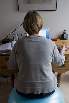 Mature woman by her desk in her home working with her laptop sitting on a pilates ball.