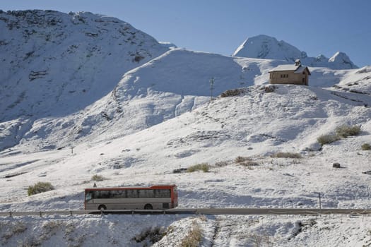 Chapel in the Dolomites in the Veneto province of Northern Italy in late September. Seen from the mountain pass Pordoi 2245 m above sea an early morning after snowfall. A public bus passing by.