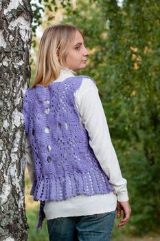 A girl in a lilac vest and white polo-neck sweater and jeans in a forest
