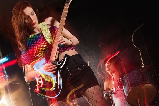 Photo of a young beautiful woman playing guitar with drummer in the background.  Motion blur and light trails from slow shutter speed and strobes. Space for text.
