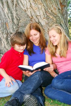 Family of mother and children reading a book under a tree in summer park