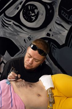 A tattoo artist applying his craft onto the abdomen of a female. (Property release supplied includes tattooists' wall mural behind him.)
