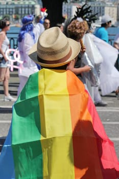 Person wearing a hat made of straw and a big rainbow flag while watching and participating at the Gaypride