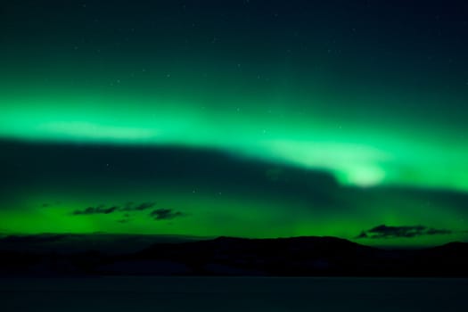Green northern lights (aurora borealis) substorm above silhouette of hills and clouds.