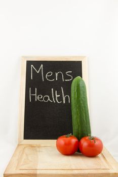 A chopping board with phallic salad, cucumber and tomato, with a chalk board in the background with the words 'mens health' all on white.