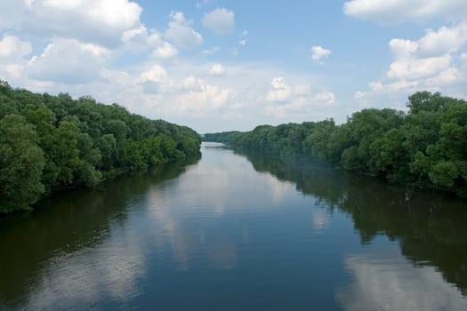 Summer. River. Blue sky and green trees are reflected in water.