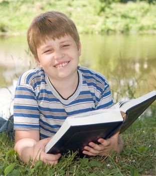 A little boy reads a big book with river at background