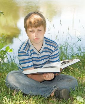 A little boy reads a big book with river at background