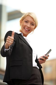 Young businesswoman looking at camera outside office building, showing thumbs up sign