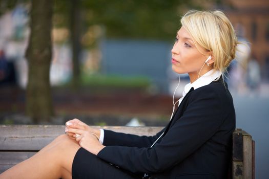 Young businesswoman wearing earphones, sitting on  bench, side view