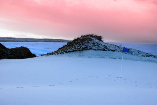 a snow covered links golf hole in ireland with blue flag and red sunset sky
