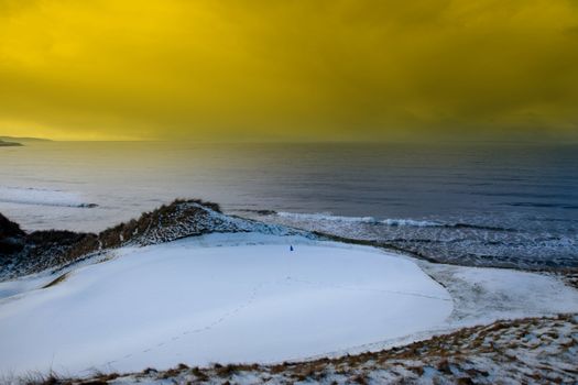 a snow covered links golf hole in ireland with blue flag and yellow sunset sky