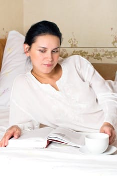 Portrait of young woman with white cup and book on bed at bedroom