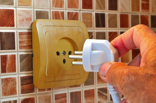 The man held the electrical plug into an outlet on a wall covered with ceramic tiles