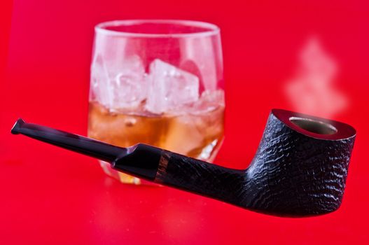 pipe for smoking tobacco and a glass of whiskey with ice photographed against a red background