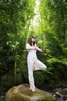 Female meditating in tropical rainforest, standing on a boulder.