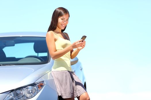 Woman standing by car sending sms text message on mobile phone. Beautiful bright summer day outdoors. Cute multiracial young lady.