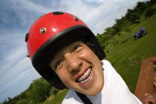 An ecstatic Asian boy with red helmet, getting ready to ride an all terrain vehicle parked in the background.
