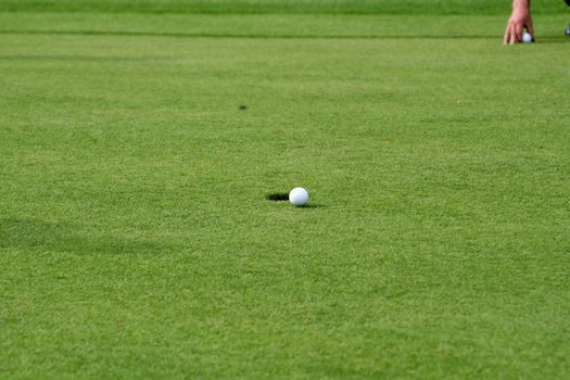Golf ball in front of the hole with a hand placing a golf ball in the upper edge