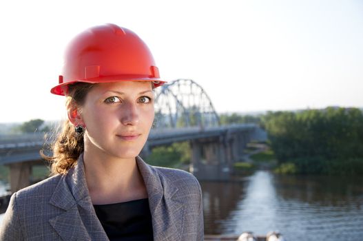Young architect-woman wearing a protective helmet standing on the bridge background is smiling
