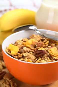 Healthy and delicious breakfast with a bowl full of wholewheat flakes mixed with banana chips, walnuts and pecan nuts surrounded by nuts and fresh banana with milk in a jug in the back (Selective Focus, Focus on the pecan nut one third into the bowl)