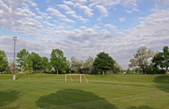 A mixed sky over an unoccupied soccer field with trees in the background.
