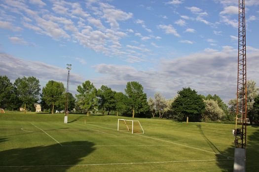 A mixed sky over an unoccupied soccer field with trees in the background.
