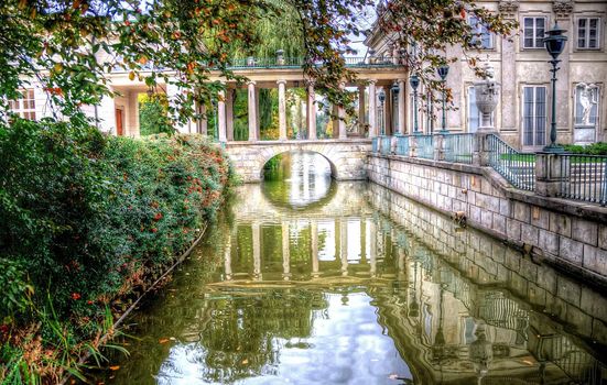 picture of lazienki park warsaw poland taken in autumn showing reflection of an arch, a historic building, lake, trees, green leaves, branches