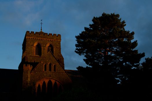 A church tower made of stone eerily glows red in the evening light next to a pine tree.