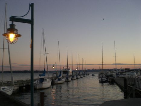 A serene sunset scene of sailboats docked at a pier in Lake Washington with the Seattle skyline on the horizon