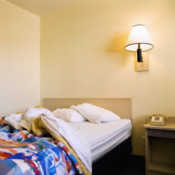 Interior shot of motel room with unmade bed and wall lamp.