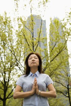 Mid-adult Asian woman standing outside with trees and skyscrapers behind her meditating.