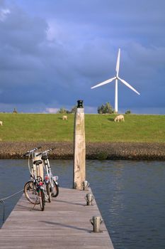 Eco-friendly means of transport and renewable energy. Bicycles above the canal and modern windmill by sunset.