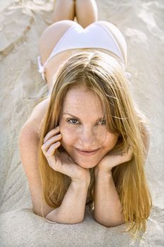 Portrait of pretty redheaded female lying in sand wearing bikini.