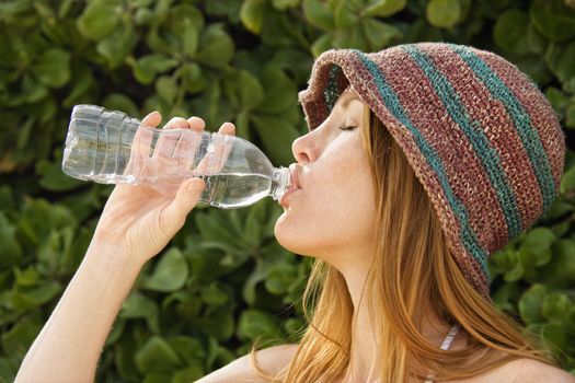 Side view of young redheaded woman wearing hat and drinking from water bottle.