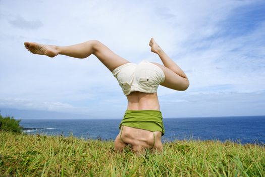 Young woman doing headstand with legs askew in grass near ocean in Maui, Hawaii.