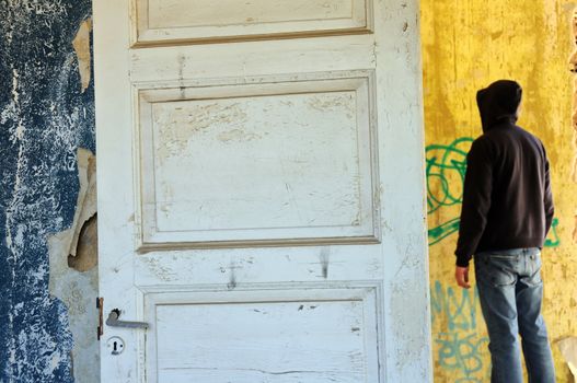 Hooded figure in abandoned house. Empty room weathered walls torn wallpaper.