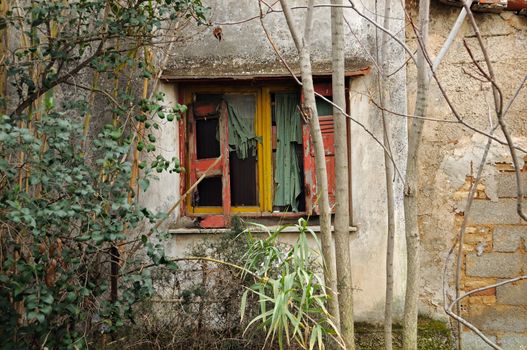 Broken window shutter and ragged drapes. Weathered abandoned house wall.