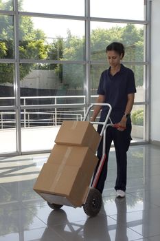 Side view of delivery woman in uniform pushing stack of cardboard boxes on dolly