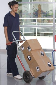 Side view of delivery woman in uniform pushing stack of cardboard boxes on dolly