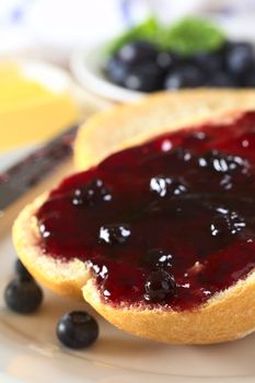 Blueberry jam on half a bun with butter and fresh blueberries in the back (Selective Focus, Focus on the front of the bun and jam)