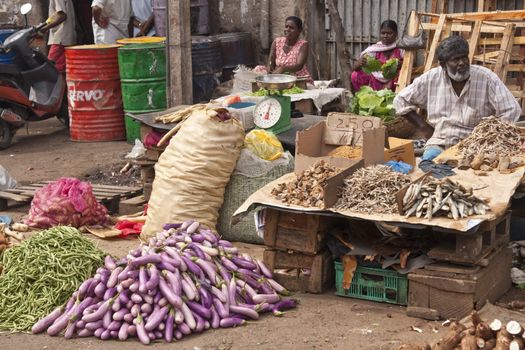 Man selling dried fish from a stall at the main food market in Colombo, Sri Lanka. Piles of fresh vegetables in the foreground.