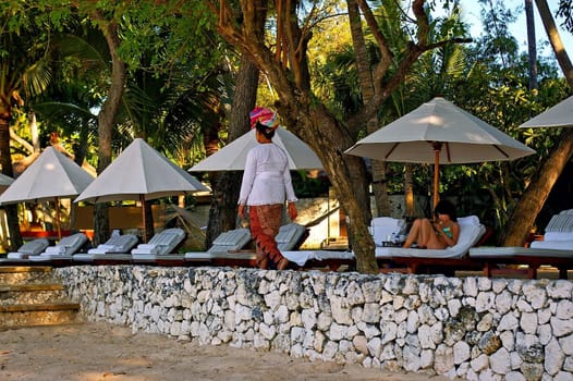 A tourist relaxing on a sun bed as a local balinese hotel worker walks past, Jimbaran beach, Bali, Indonesia