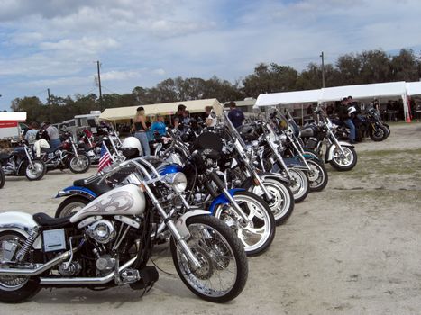 A whole range of motorcycles are displayed side by side while the owners socialize in the background during BikeWeek.