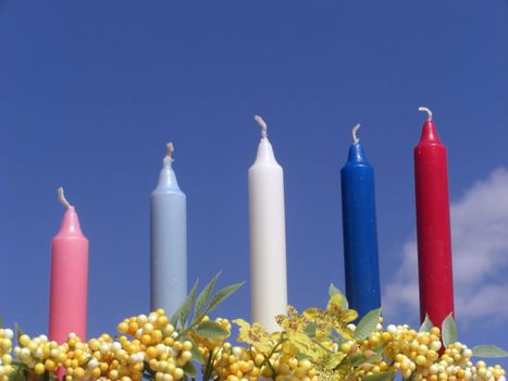 A set of five colorful candles are set in between yellow flowers and against a blue sky.
