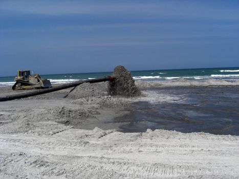 A big pipe is sucking beach sand that had eroded into the ocean during hurricane season, while a bulldozer pushes and levels it to reclaim the beach.