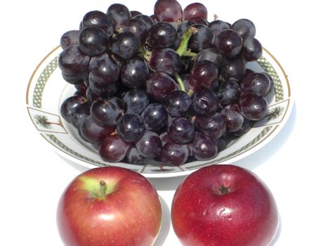 A bowl is full of nutritious, fresh and juicy looking black grapes and two red apples on the side.
