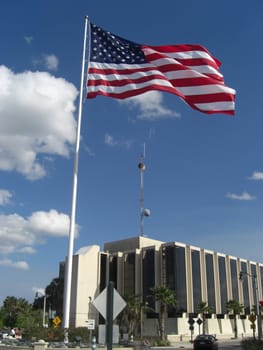 A flag is standing way above the ground and is towering over a building with a satellite on the roof.