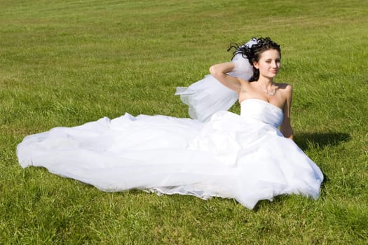 a bride in a beautiful long white dress sits on the green grass and smiles