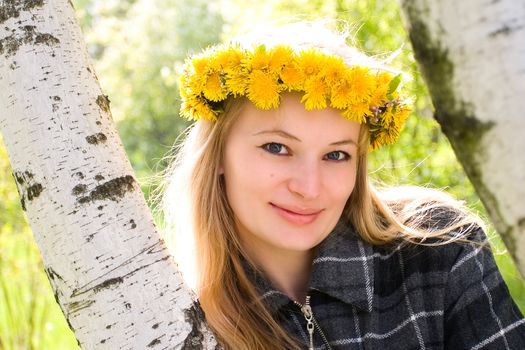 The young girl in a wreath from dandelions stand near a birch